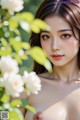 A woman with long brown hair and makeup, surrounded by greenery and white flowers.