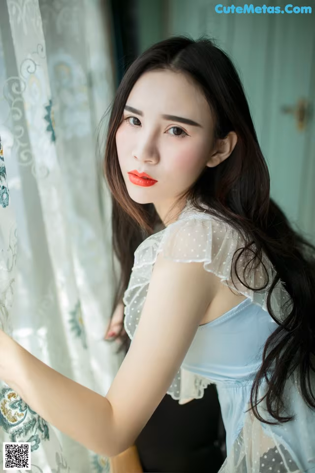 A woman in a blue dress leaning against a curtain.